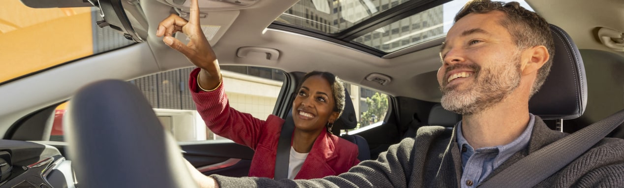 A woman pressing the OnStar button as the driver smiles upon inside their vehicle.